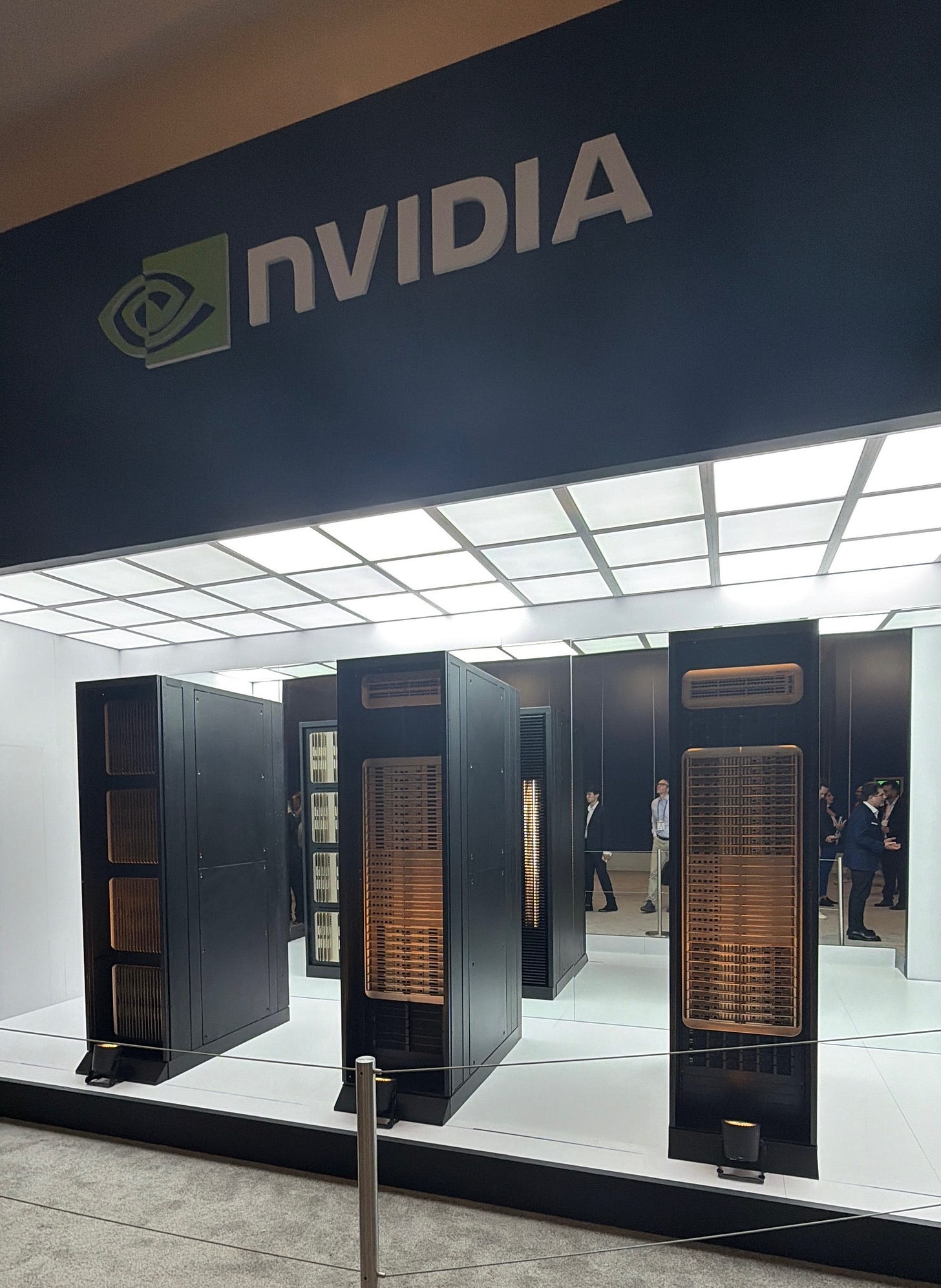 A display of NVIDIA’s Vera Rubin server racks at CES, shown in a brightly lit booth with large NVIDIA branding above. Several black and copper cooling-heavy units stand on a raised platform with attendees visible in the background. A display of NVIDIA’s Vera Rubin server racks at CES, shown in a brightly lit booth with large NVIDIA branding above. Several black and copper cooling-heavy units stand on a raised platform with attendees visible in the background.