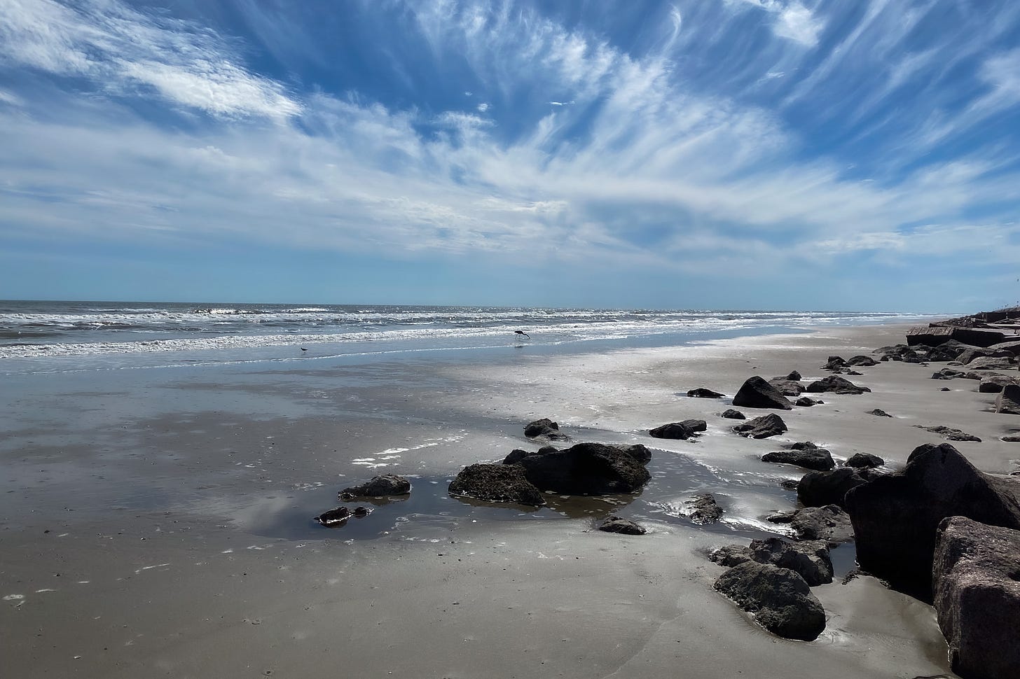The beach with large boulders on the right. the waves crashing on the shore on the left. and a streak of white clouds in a blue sky seeming to mirror the angles of the shoreline and rocks.