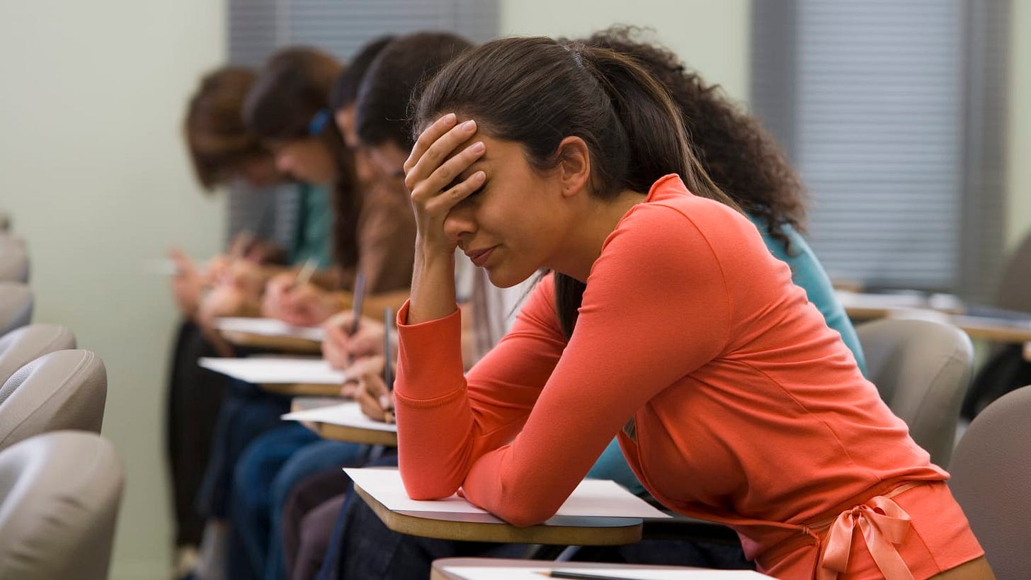 University student wearing a salmon colored top and pony tail sits at desk while taking test with head in her hand. University student wearing a salmon colored top and pony tail sits at desk while taking test with head in her hand.
