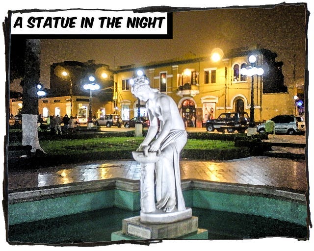 A white statue standing in a fountain in front of several buildings. It’s a night scene and lights are reflected on the slick pavement. The caption reads, “A statue in the night.”