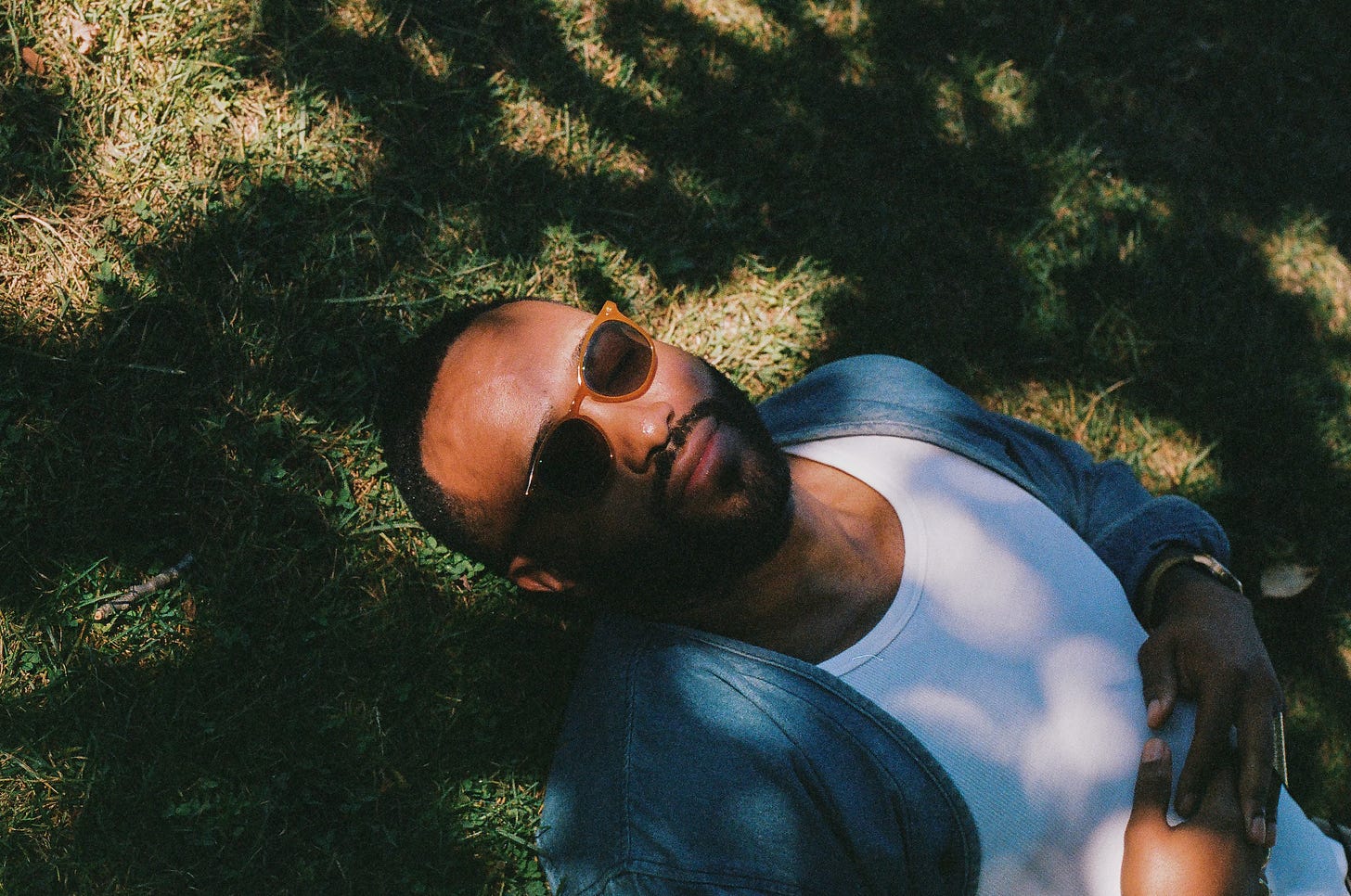 A man lying in the grass in soft sunlight with sunglasses on, resting quietly in a moment of stillness.