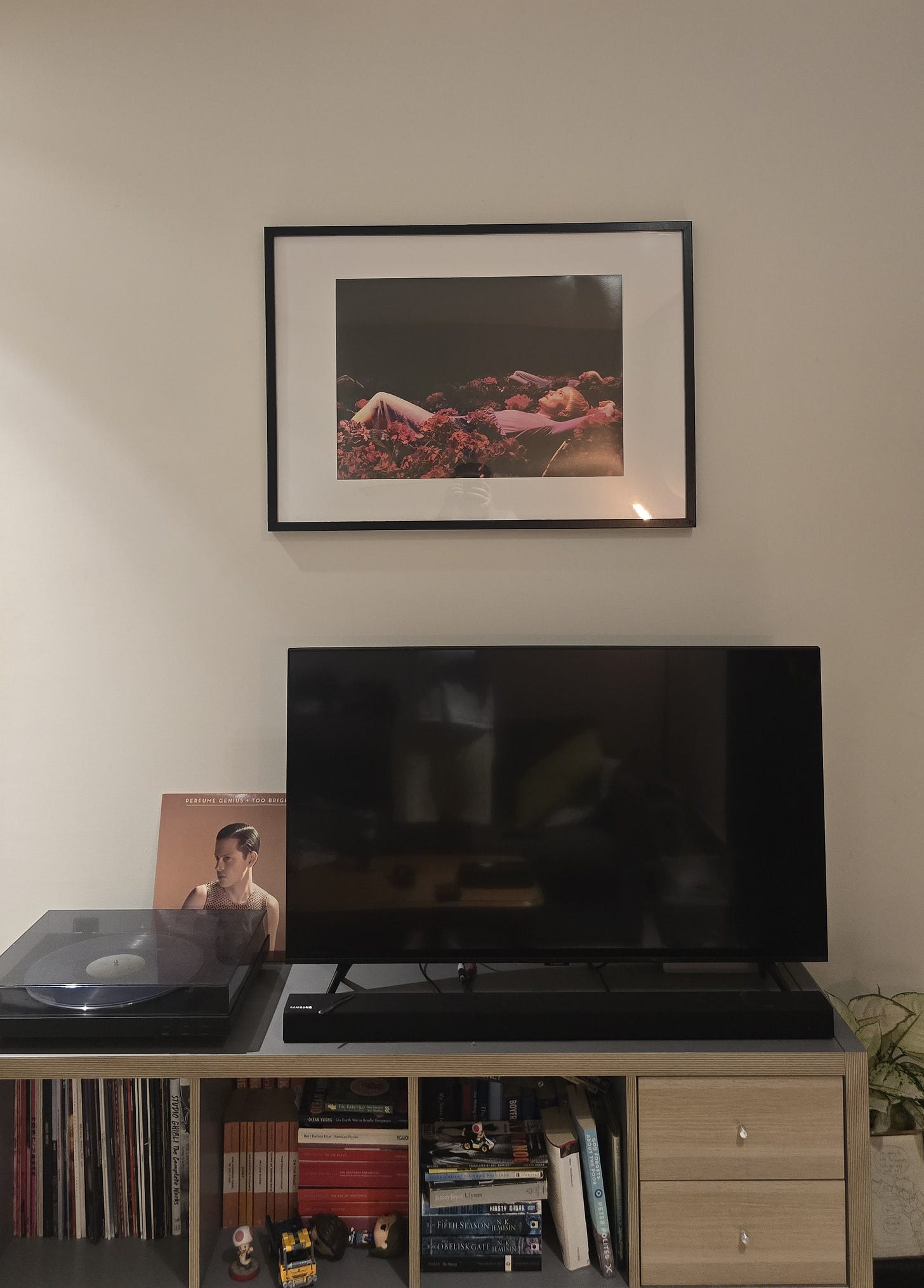 A photo of the wall of my living room. There's a TV on an Ikea cabinet full of books and some Gilmore Girls funko pops and a little Toad from Mario kart. There's a bunch of books and on top a record player with the 10th anniversary vinyl press of 'Too Bright' by Perfume Genuis on it, the cover sitting against the wall. The TV is off, and above it on the wall there's a framed production still from A Room of One's Own, where a person with short blonde hair lies on their back surrounded by roses on an otherwise empty background.
