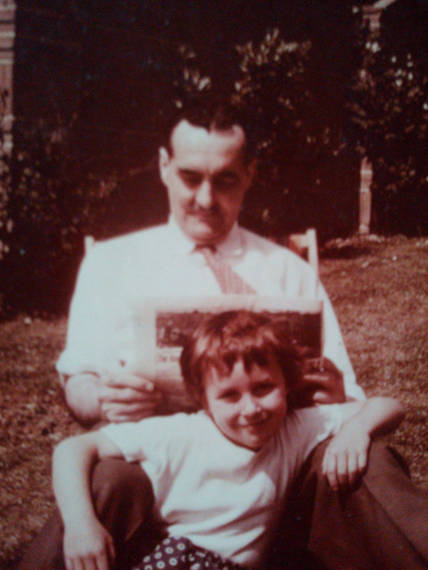 A man with a mostache sits in a deck chair reading while his young daughter leans up against his knees. She looks out at us smiling at the photographer.