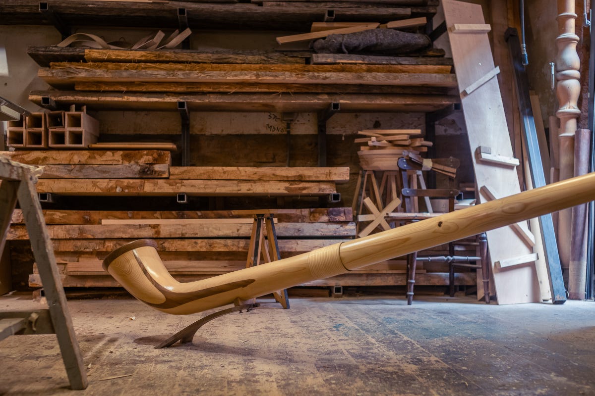 A wooden alphorn resting on a stand in a rustic woodworking studio filled with lumber and tools.