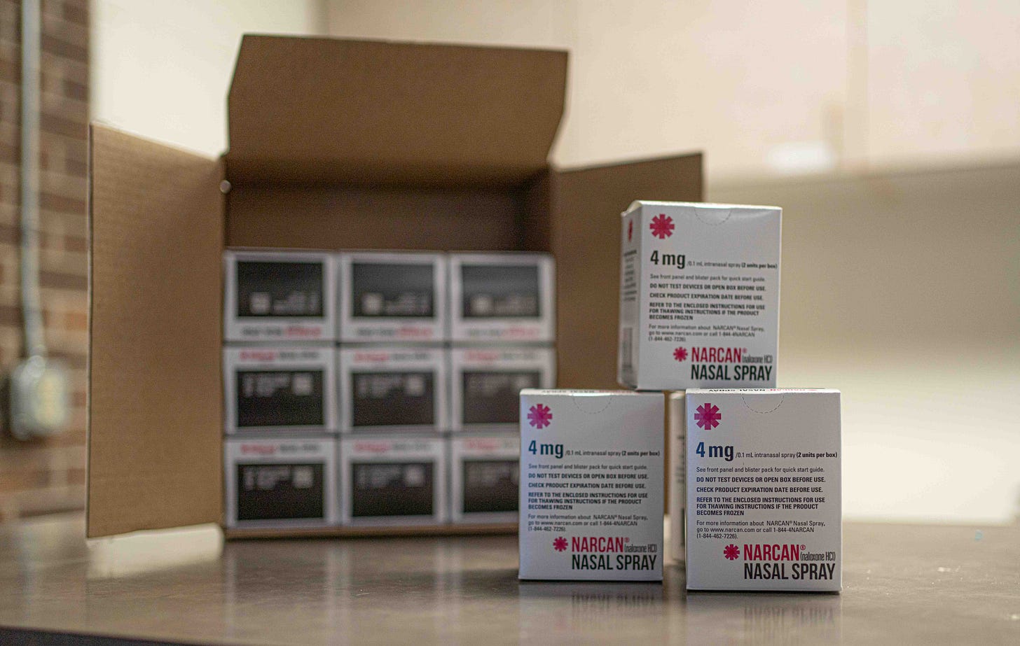 Three stacked boxes of "4mg Narcan Nasal Spray" are stacked in front of a larger shipping box containing even more nasal sprays. Three stacked boxes of "4mg Narcan Nasal Spray" are stacked in front of a larger shipping box containing even more nasal sprays.