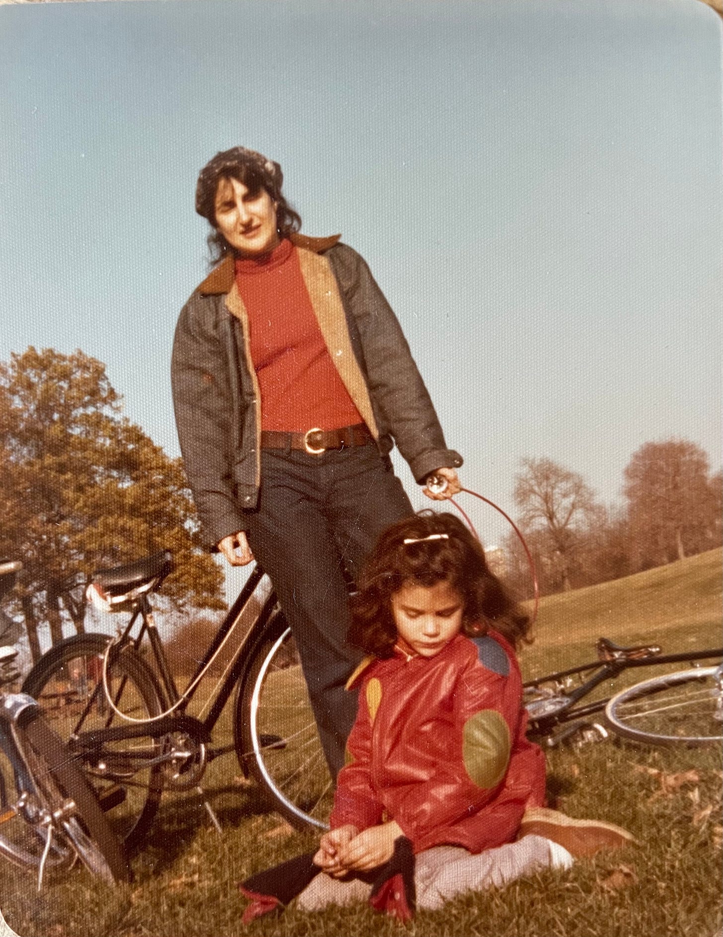 A woman in her 40s stands backdropped with blue sky and trees above a girl seated in the grass, bicycles behind them. The child looks down plaintively as the woman smiles.