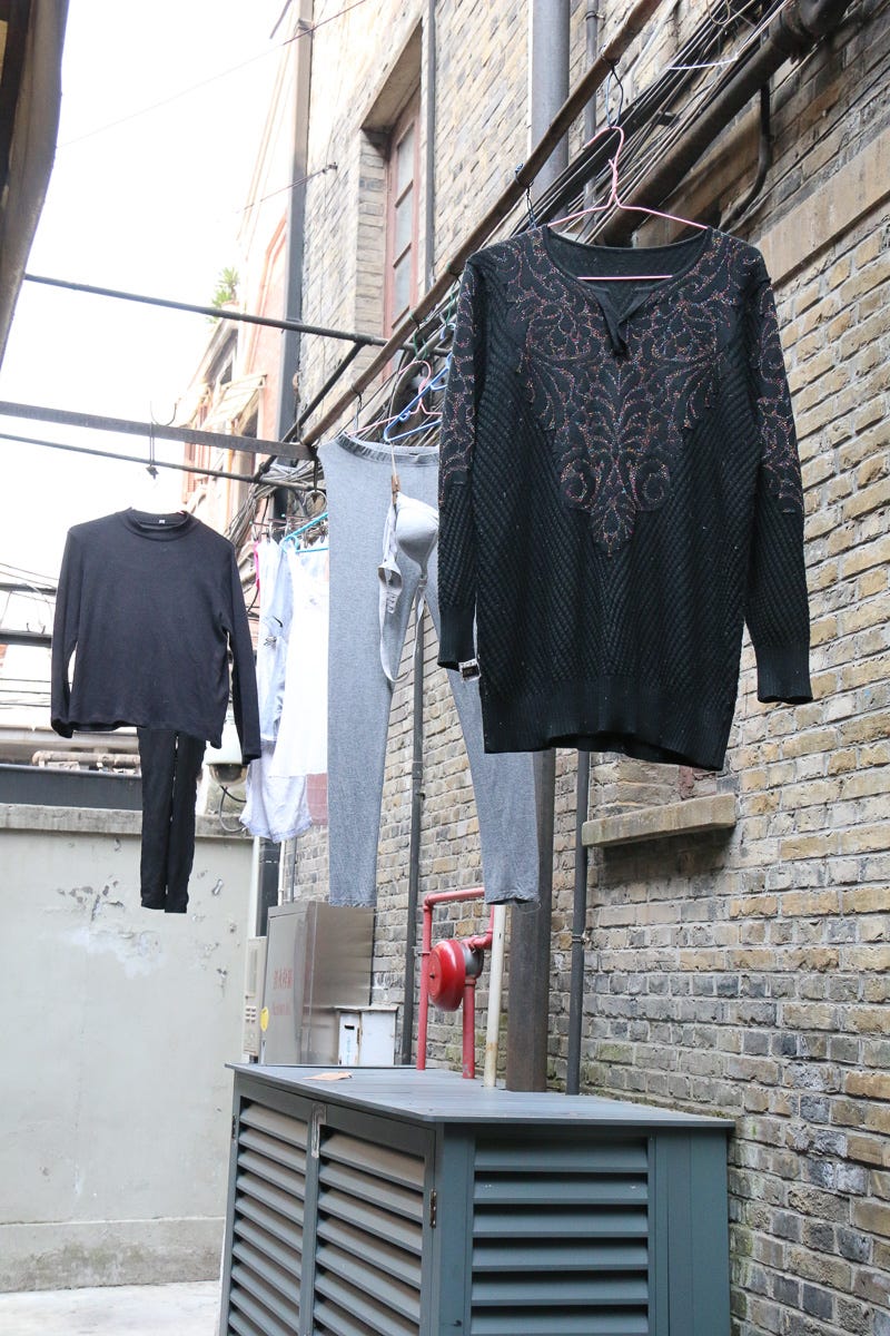 Black and gray clothing dries along a narrow stone alleyway. A quiet, hidden domestic moment amidst tight Shanghai lanes.