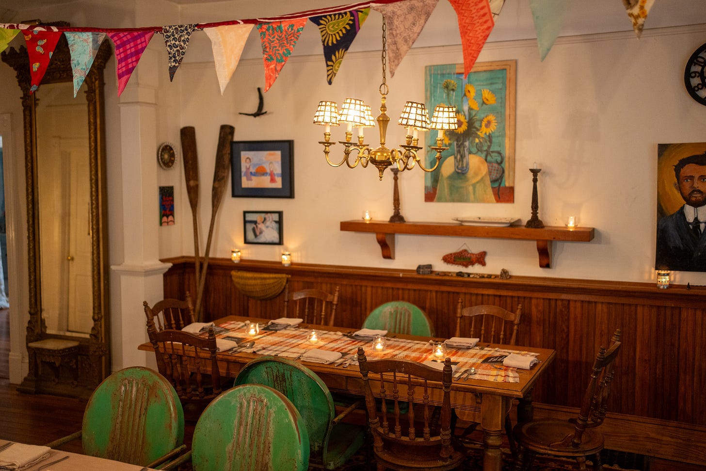 A dimly lit dining room with wood-paneled walls, a long wooden table set for dinner with candles, green wooden chairs, and colorful fabric bunting hung from the ceiling.
