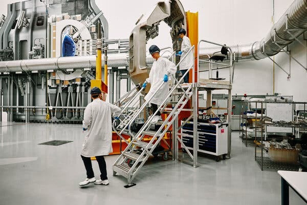 Three people in white lab coats are near a set of metal stairs in a large open room with pipes and other machinery. 
