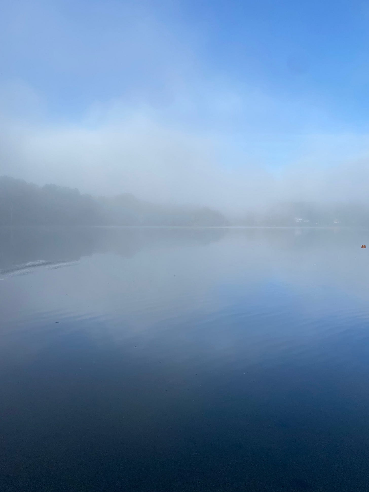View of a misty lake; the water and line of hills on the horizon are vague and blurry behind the fog.