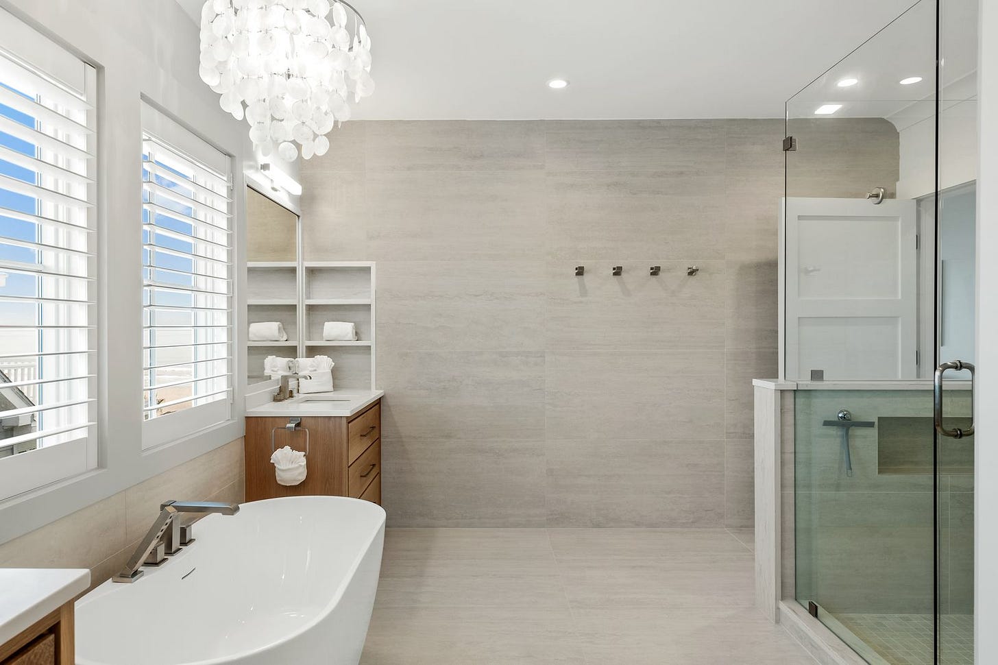A light-filled bathroom with large-format tile running floor to ceiling on every wall, a freestanding white soaking tub, a wood vanity, a glass-enclosed shower, and plantation shutters on the windows. A light-filled bathroom with large-format tile running floor to ceiling on every wall, a freestanding white soaking tub, a wood vanity, a glass-enclosed shower, and plantation shutters on the windows.