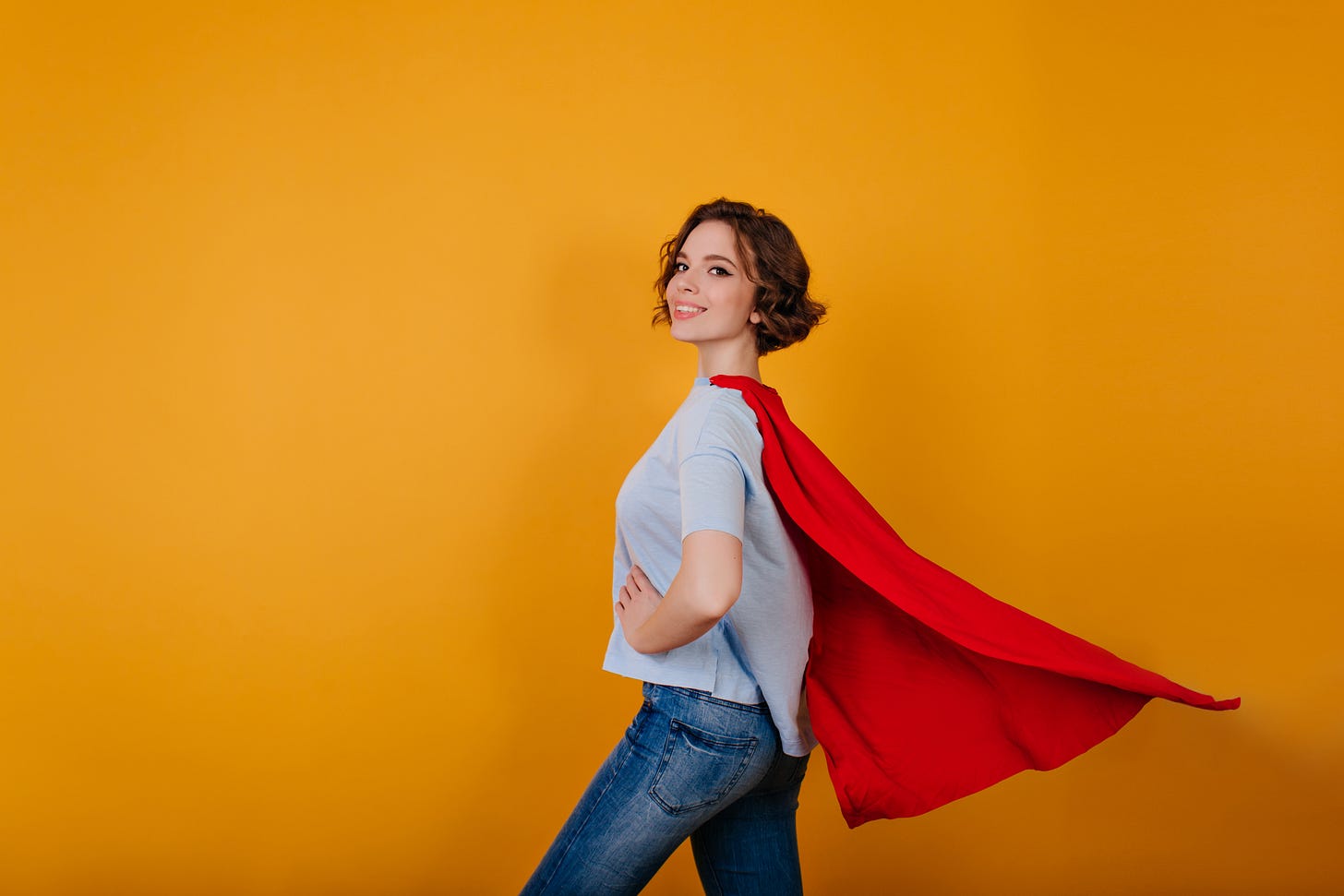 A woman with short wavy hair wearing jeans, a t-shirt and a red cape, standing sideways against a yellow background A woman with short wavy hair wearing jeans, a t-shirt and a red cape, standing sideways against a yellow background