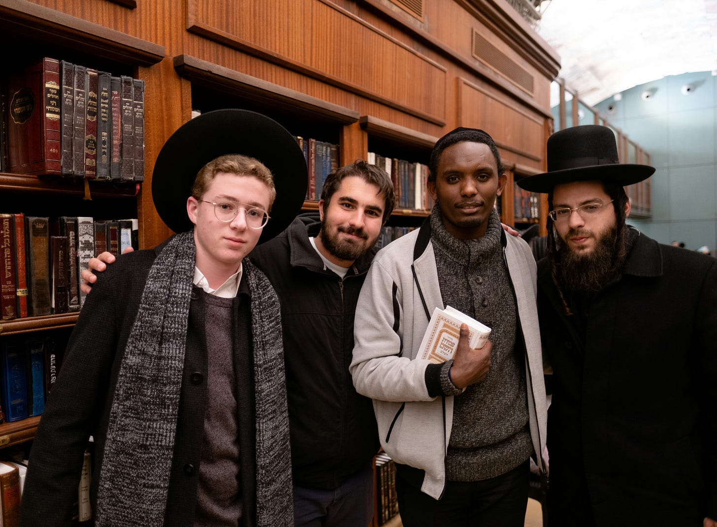 Photo of 4 young men in front of a wall of Hebrew books: on ultraOrthodox, one Ethiopian, one who might be secular, and one dressed between modern Orthodox and Charedi