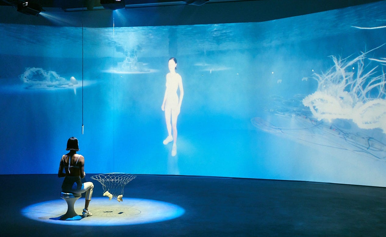 An artist sits on a stool under a bright spotligth in front of an interactive virtual reality environment in which visitors converse with an AI water nymph modelled on the artist's body.