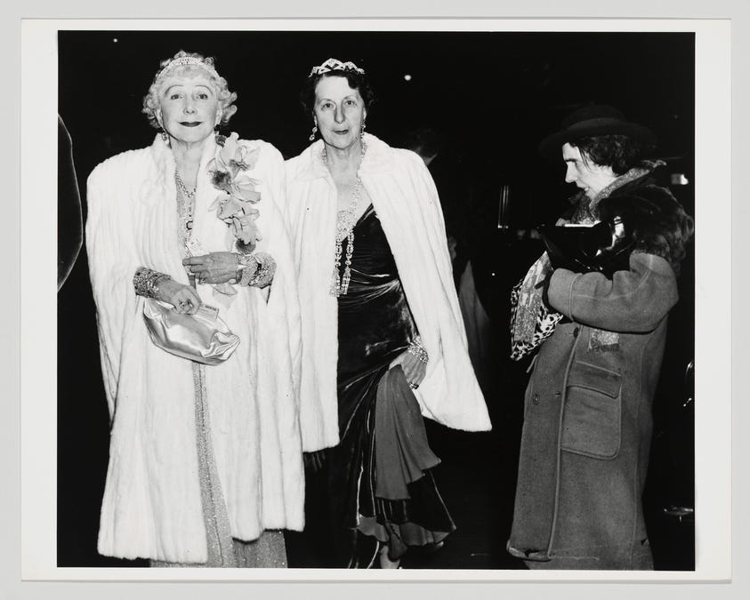 Two elegantly dressed women in fur coats and tiaras at a social event, with a third woman in a coat looking on.
