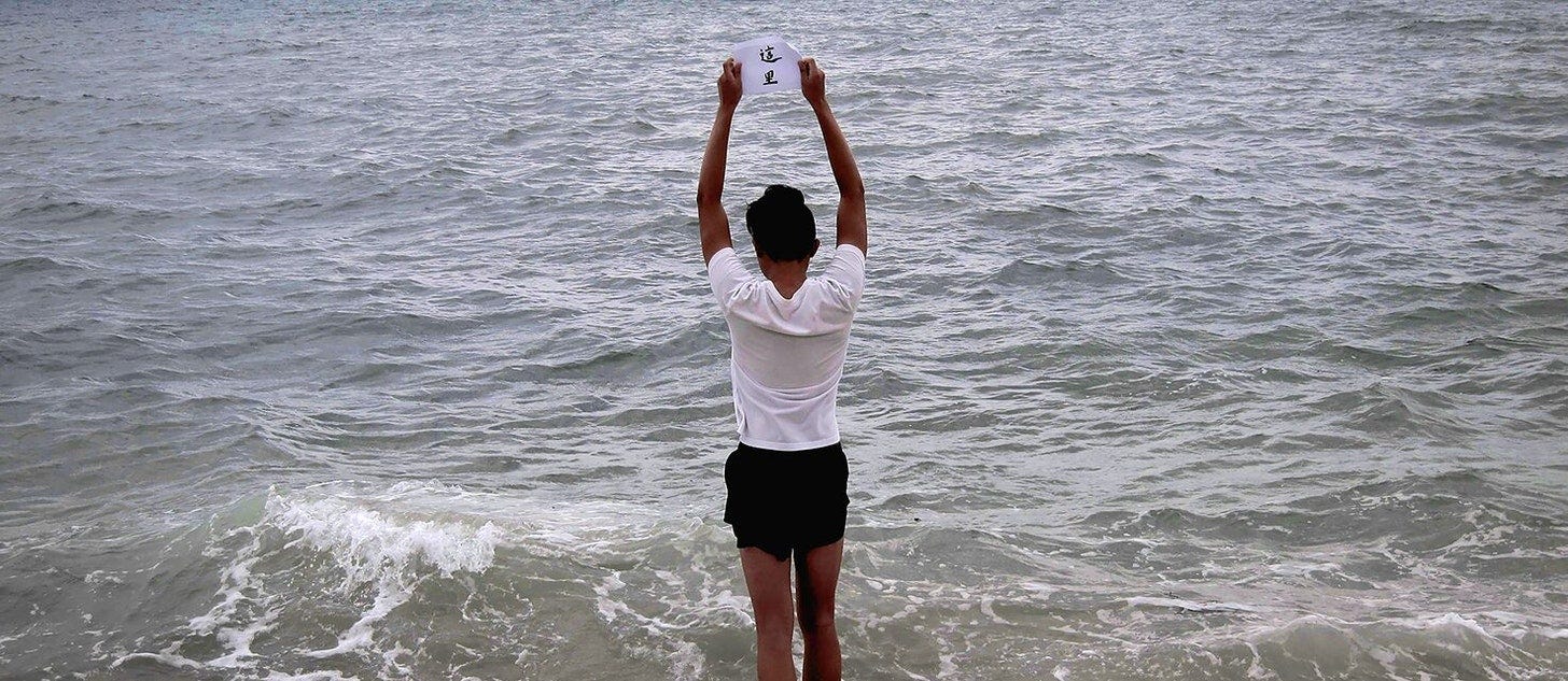 jee chan standing in the waves at East Coast Park, holding up a piece of calligraphy written by their grandmother/婆婆 that reads 這里, meaning “here” jee chan standing in the waves at East Coast Park, holding up a piece of calligraphy written by their grandmother/婆婆 that reads 這里, meaning “here”