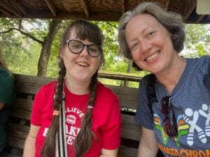 Two smiling people sit together on a wooden bench outdoors. The girl on the left, wearing glasses, has long brown hair in braids and a red T-shirt. The woman on the right has short light brown hair, is wearing a gray graphic T-shirt, and has sunglasses hanging from the collar. Green trees and wooden beams of the shelter are visible in the background.
