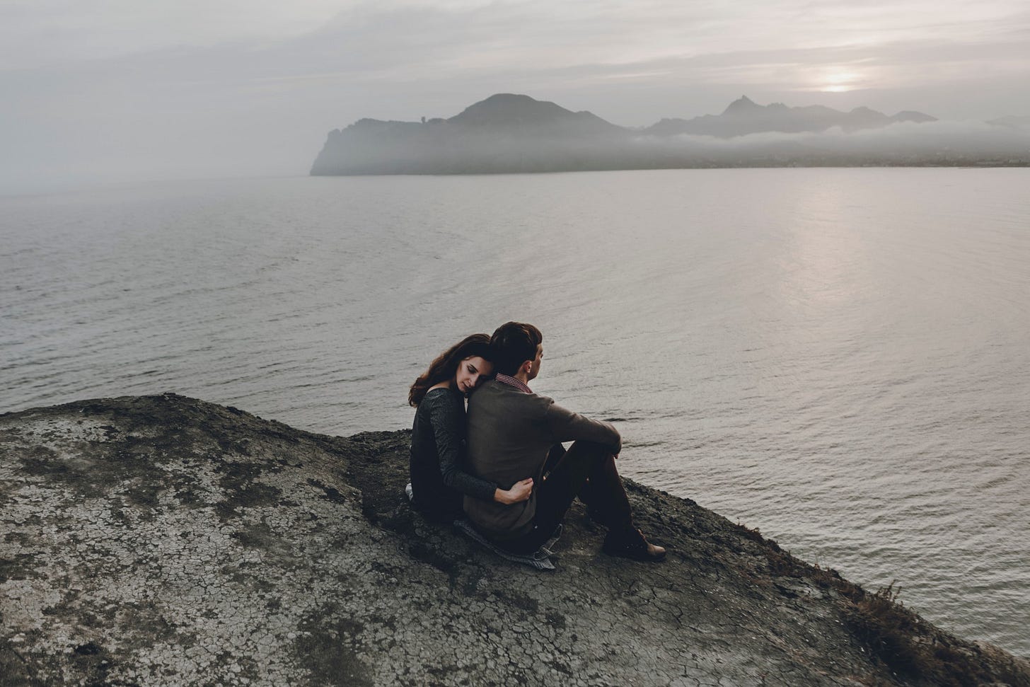 A couple embraces on a cliff overlooking a grey and cloudy sea.
