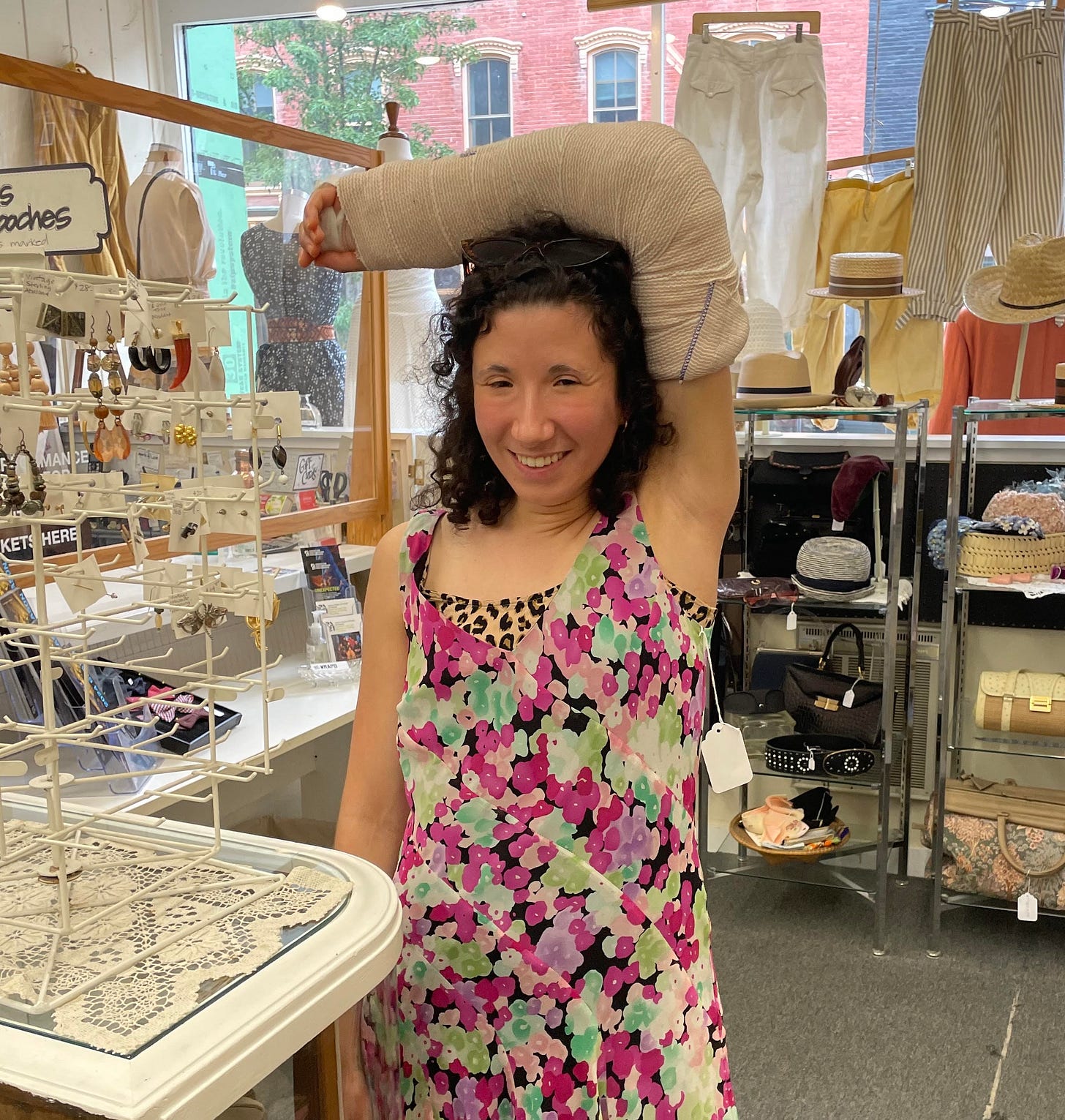 White woman with curly hair wearing a floral dress with her left arm in a giant cast balanced on her head. She is standing in a shop surrounded by knick-knacks and jewelry.