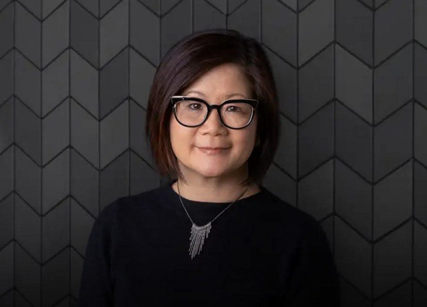 Gloria Chen, Adobe’s Chief People Officer, standing in front of a dark, geometric-patterned wall. She is wearing glasses, a black top, and a pendant necklace.