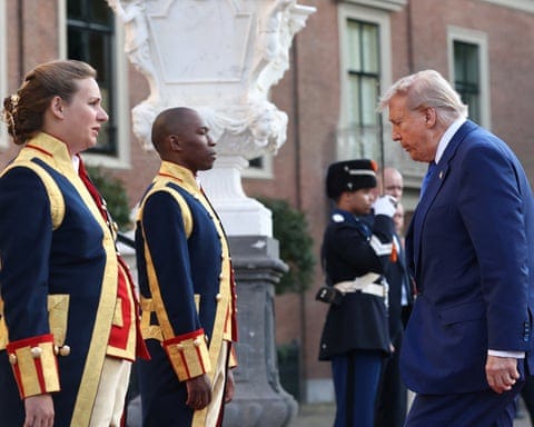 Trump, wearing a blue suit, walks to the palace doorway; guards in dark blue, gold and red uniforms stand to either side.