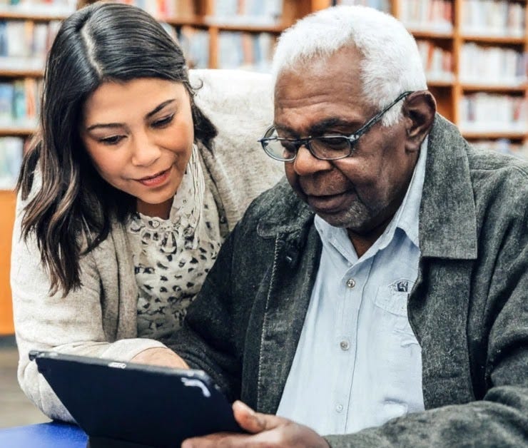 A young nurse helps an older man with his tablet A senior man with a long-term illness uses a tablet, while a smiling female healthcare professional stands beside him.