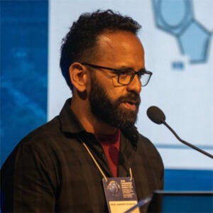 A imagem mostra um homem falando em um microfone durante uma apresentação ou palestra.Ele está de perfil, levemente virado para a esquerda, olhando para frente enquanto fala. O homem tem cabelo curto e encaracolado, barba cheia e escura e usa óculos de armação escura. Ele veste uma camisa escura, aberta por cima de uma camiseta de tom vinho ou vermelho escuro. No pescoço, há um cordão com crachá, o que sugere que ele está participando de um evento, congresso ou conferência. À frente dele há um microfone em um suporte, posicionado próximo à boca. No fundo da imagem aparece uma tela de apresentação, com gráficos ou figuras desfocadas, indicando que ele provavelmente está explicando algum conteúdo para um público. A iluminação é típica de auditórios ou eventos acadêmicos, com foco na pessoa que está falando.
