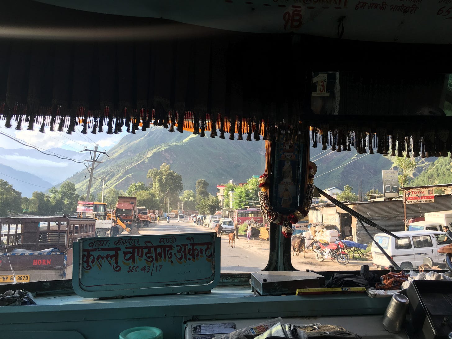The view from the front window of a bus. Green hills and clouds are in the distance. Cars, buildings, and cows are seen in the foreground.