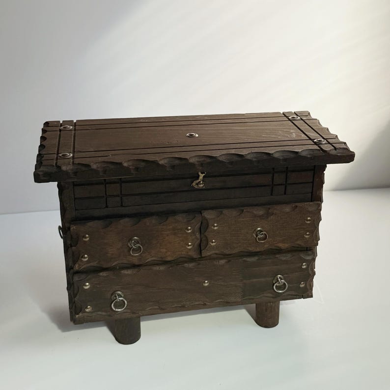 May include: A dark brown wooden chest with a hinged lid and two drawers. The chest has decorative metal accents and ring pulls on the drawers. It is supported by four short, rounded legs.