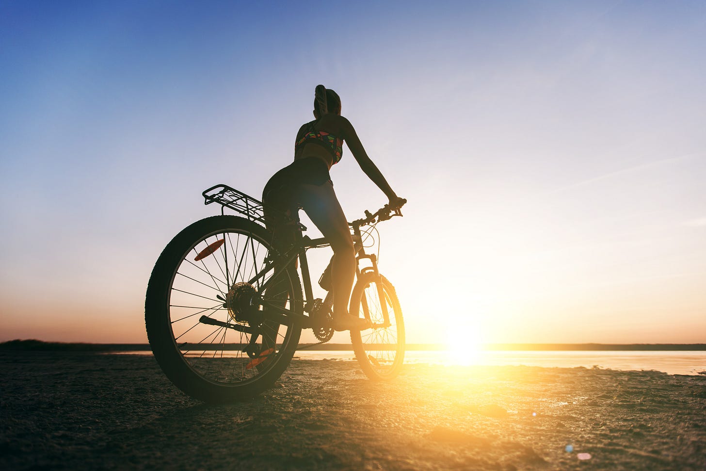 Silhouette of a cyclist at sunrise by the water, sunlight flaring through the wheel.