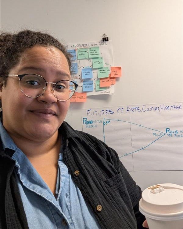 A portrait photo of Danielle Nadine Pierre, a Black-Canadian-Caribbean woman, wearing glasses, a blue shirt under a black shirt and holding a coffee. In the background are drawings and post-it notes.