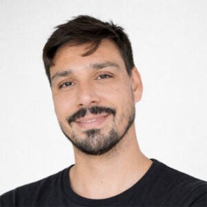 Homem de cabelos escuros e barba curta, com um sorriso leve, usando camiseta preta, diante de fundo claro. Homem de cabelos escuros e barba curta, com um sorriso leve, usando camiseta preta, diante de fundo claro.