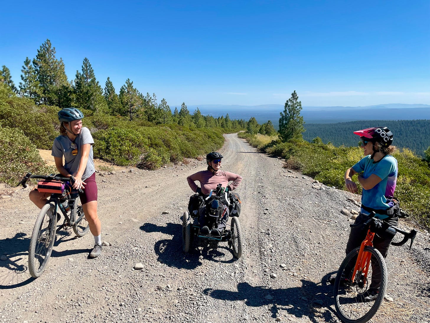 3 gals on bikes — 2 gravel bikes and 1 handcycle bike. On a gravel road in the mountains