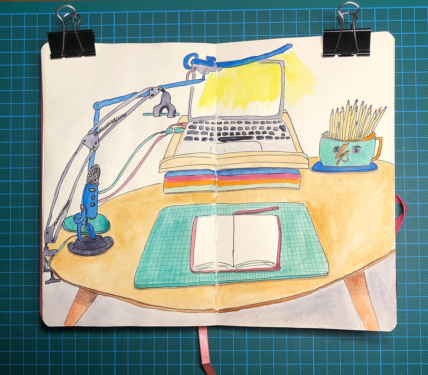 Photo d'une double-page de mon sketchbook où j'ai peins mon ordinateur, ma lampe, un mug avec des crayons de couleurs, la bras qui tient mon téléphone portable, le micro et mon sketchbook posé sur mon tapis de découpe.