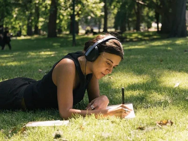 This may contain: a woman laying in the grass writing on her notebook with headphones around her ears