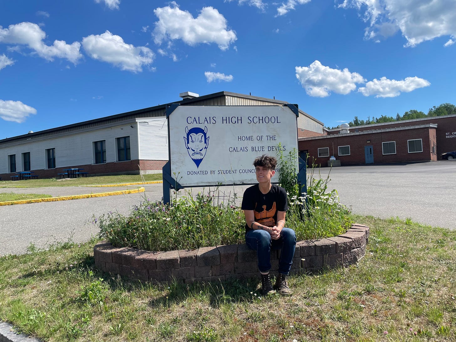 Andrea, in 2021, poses in front of their old high school, with short hair, tattoos, and boots.