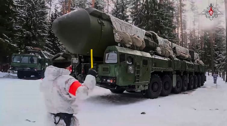 a soldier in a white uniform guides a large wheeled vehicle carrying a large missile over snow-covered ground a soldier in a white uniform guides a large wheeled vehicle carrying a large missile over snow-covered ground