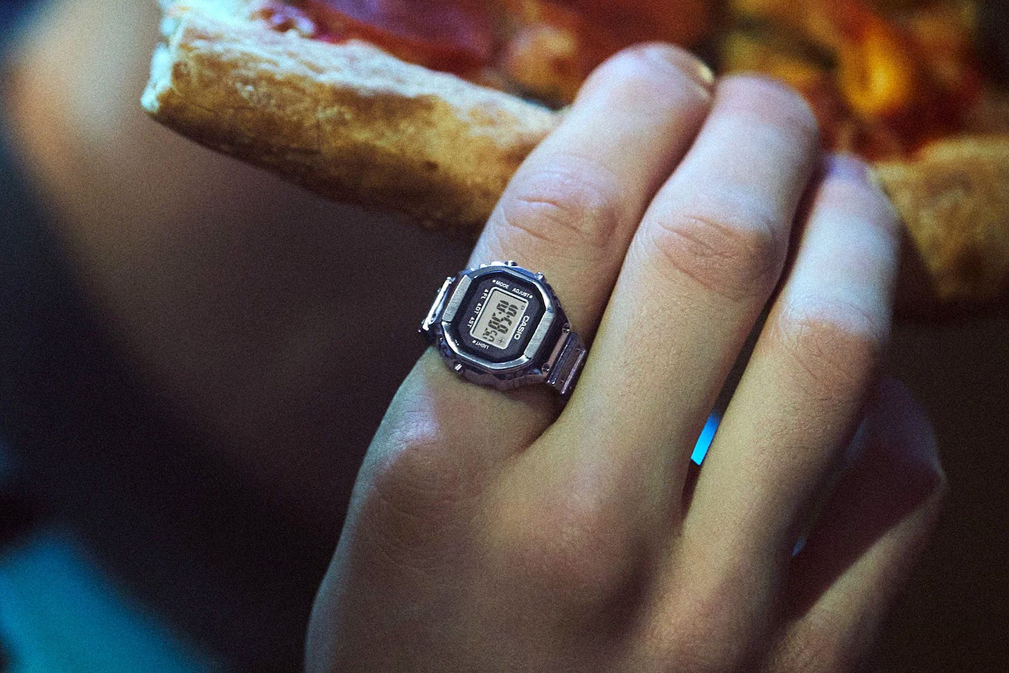 A person holding a slice of pizza while wearing Casio’s functional digital watch ring.
