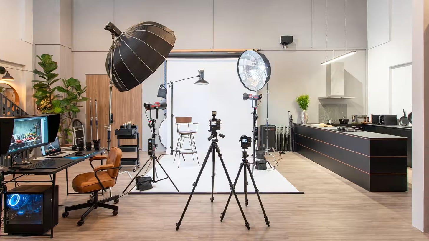 Complete professional photography studio featuring multiple camera setups on tripods facing a white cyclorama backdrop. The versatile space includes professional lighting with a large softbox and beauty dish, an editing workstation with monitor displaying an image, and modern kitchen area. Green plants add warmth to this multi-purpose creative space.