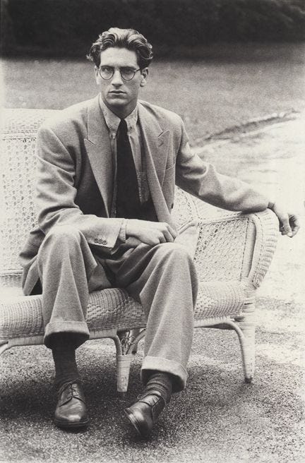 This may contain: black and white photograph of a man in suit sitting on a wicker chair with his legs crossed