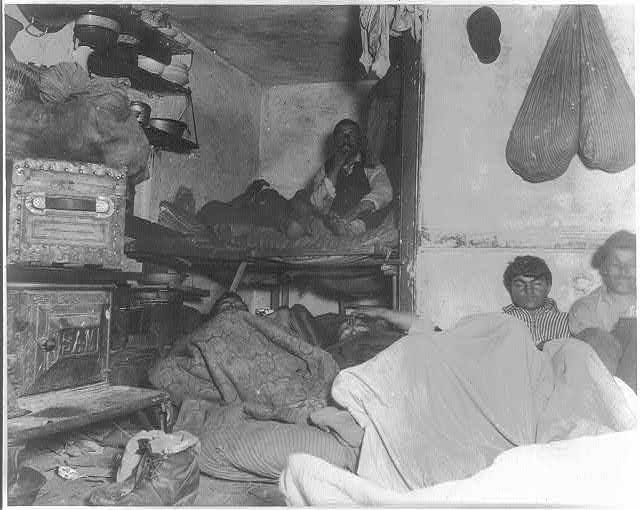 Flash photograph of a cramped Bayard Street lodging room where several adults sleep on bunks and the floor among clothing and household objects, circa 1890. Flash photograph of a cramped Bayard Street lodging room where several adults sleep on bunks and the floor among clothing and household objects, circa 1890.