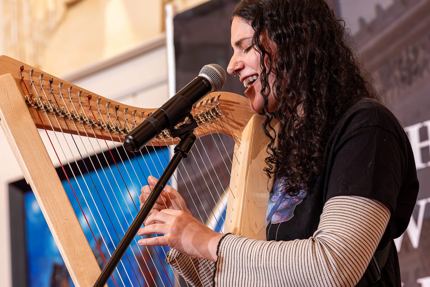 Performer: Sara Henya plucking her harp strings as she sings into the microphone Performer: Sara Henya plucking her harp strings as she sings into the microphone