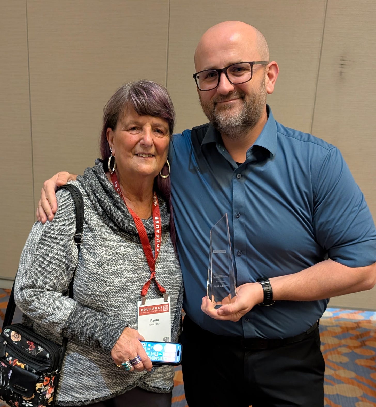 An older woman and a bald man with glasses stand with their arms around each other, smiling at the camera in a conference venue. The woman wears a gray sweater and red EDUCAUSE lanyard, while the man wears a blue shirt and holds a tall, angular glass award between them.