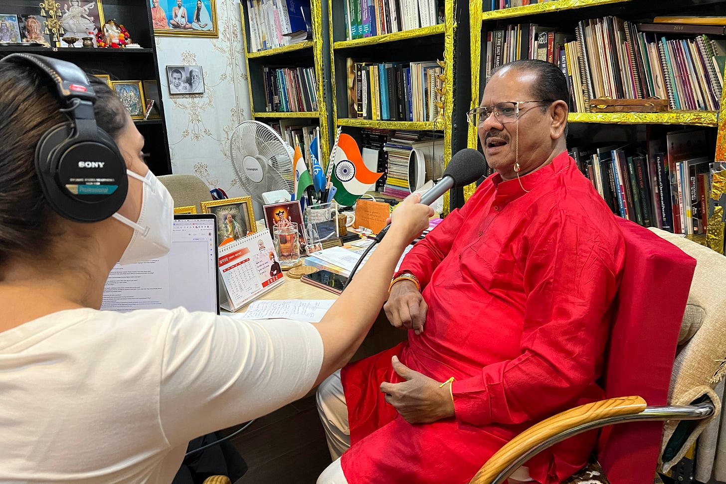 Chandrakant Pandav, wearing a bright red outfit and glasses, off of which hangs a small gold chain that links around the back of his neck, sits in his office as he is interviewed by Céline Gounder. She is to his left, holding a microphone to him as he speaks. She wears professional audio-recording headphones and a KN95 mask.