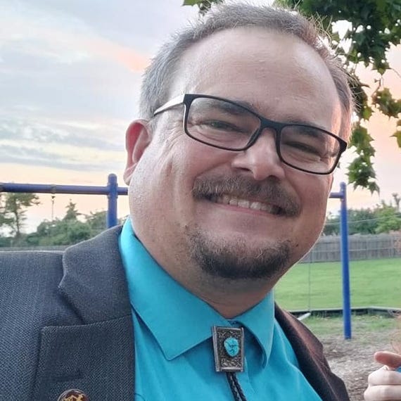 James Branum wears glasses and has a beard and mustache. He wears a turquoisse shirt with a bolo.