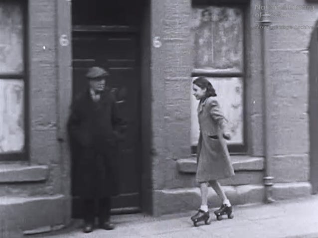 Siyah-beyaz bir fotoğraf. Biz kız patenle bir binanın önünden kayıyor. Bir kapının önünde durmuş bir adam da ona bakıyor.