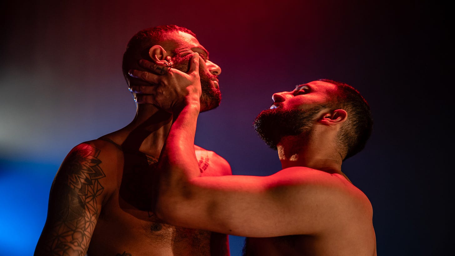 A pair of bearded and tattooed South Asian men embrace, one awkwardly clutching the other's face.