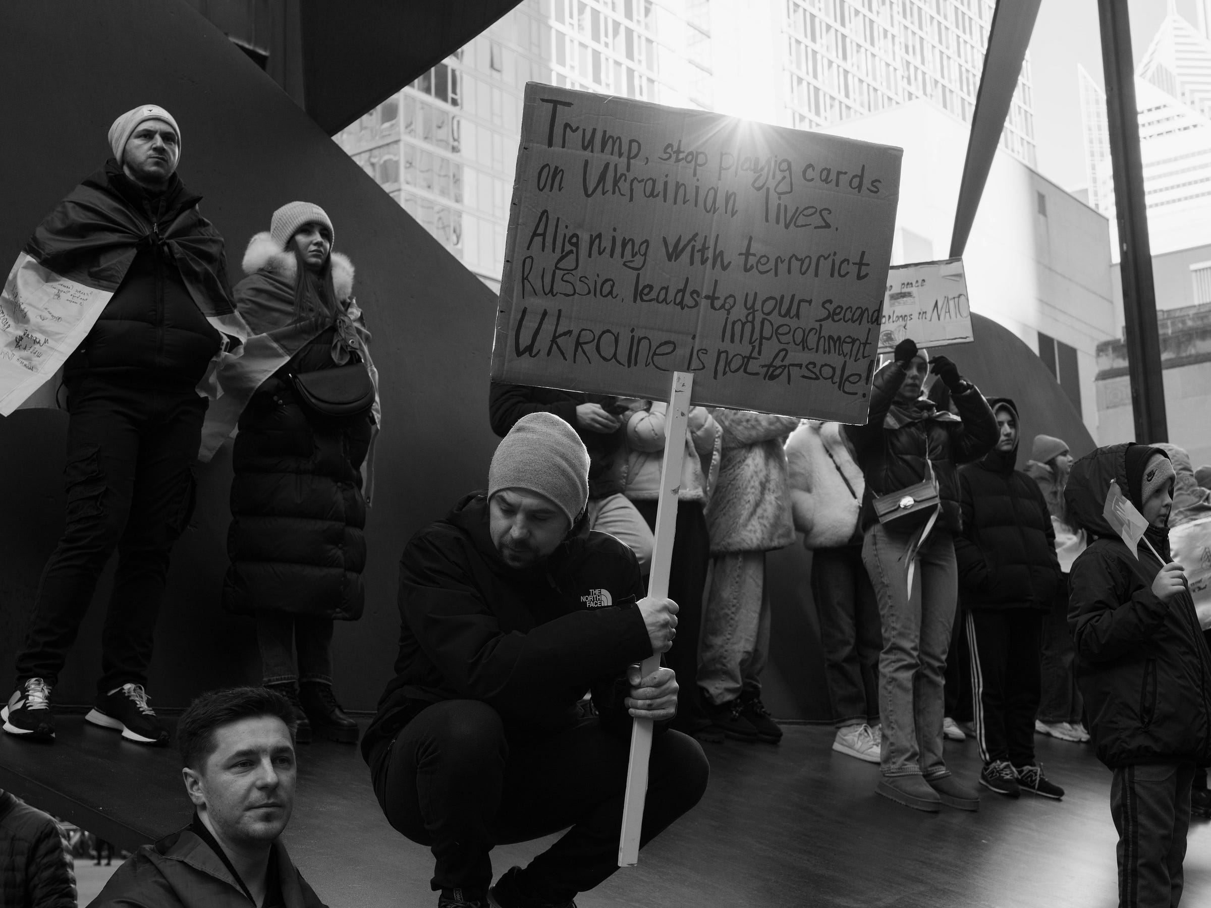 A black and white photograph captures a group of protesters gathered around a large public sculpture in an urban setting. A man crouches in the foreground, holding a sign that reads, "Trump, stop playing cards on Ukrainian lives. Aligning with terrorist Russia leads to your second impeachment. Ukraine is not for sale!" Other demonstrators, wrapped in Ukrainian flags, stand on the sculpture, looking solemn and determined. In the background, more protesters hold signs and flags, while city skyscrapers rise above them. A black and white photograph captures a group of protesters gathered around a large public sculpture in an urban setting. A man crouches in the foreground, holding a sign that reads, "Trump, stop playing cards on Ukrainian lives. Aligning with terrorist Russia leads to your second impeachment. Ukraine is not for sale!" Other demonstrators, wrapped in Ukrainian flags, stand on the sculpture, looking solemn and determined. In the background, more protesters hold signs and flags, while city skyscrapers rise above them.
