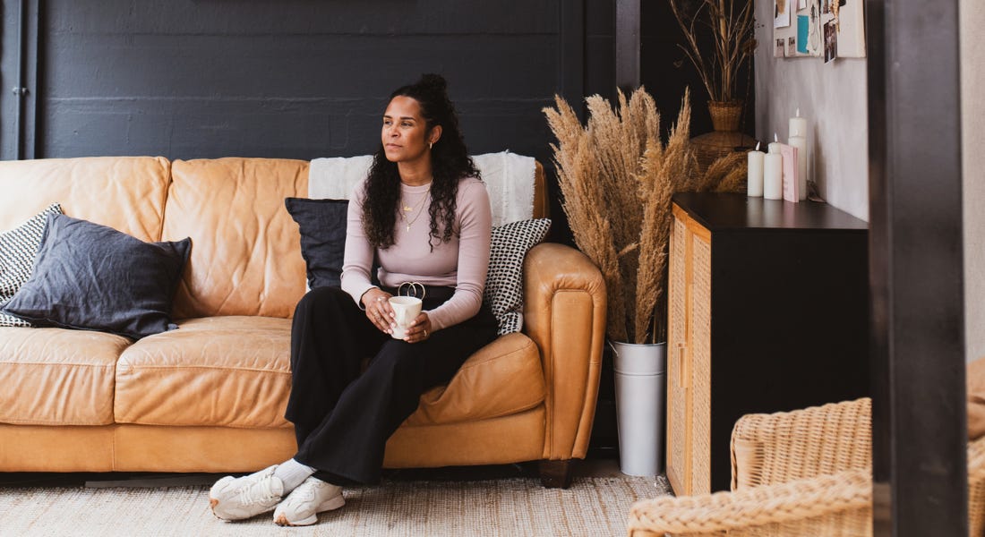 Tarnya Bruce sitting on a brown leather sofa with a black wall behind her and dried flowers next to her Tarnya Bruce sitting on a brown leather sofa with a black wall behind her and dried flowers next to her
