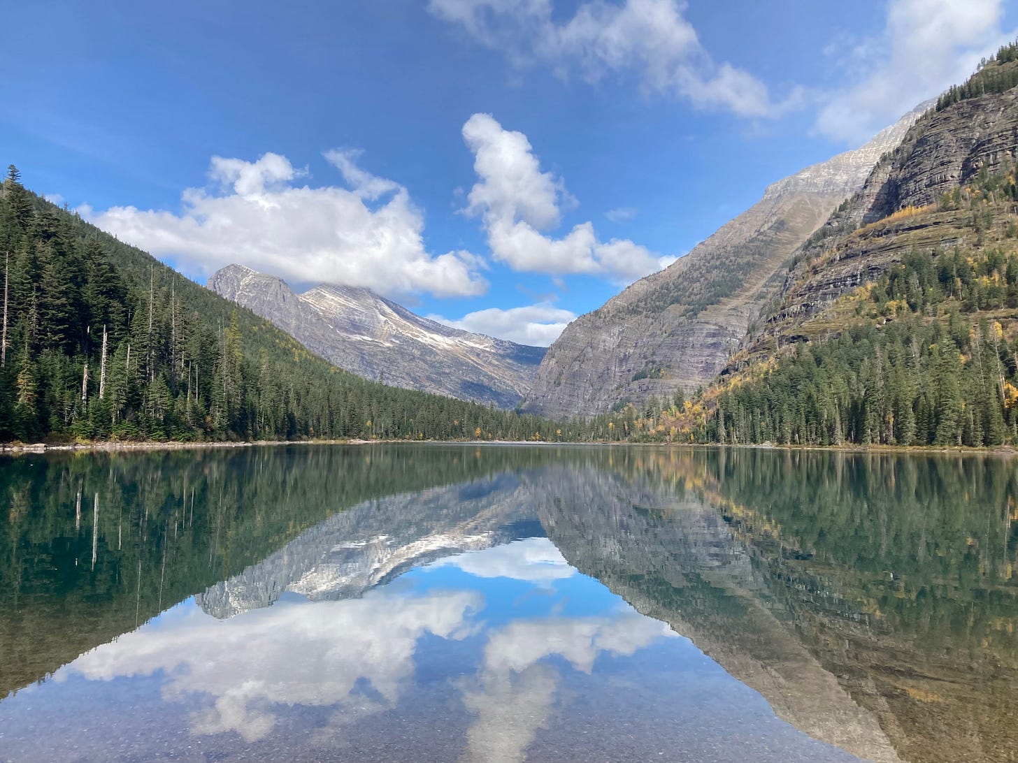 landscape view of a calm lake mirroring mountains with green pine forests and snowy tops and blue sky with clouds landscape view of a calm lake mirroring mountains with green pine forests and snowy tops and blue sky with clouds